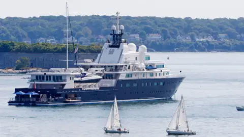 Getty Images Le Grand Bleu, a large yacht with a navy blue keel and white upper decks, with parasols visible on the large swim platform at the rear, anchored near Portland in Maine, USA, with small sailing boats passing by.